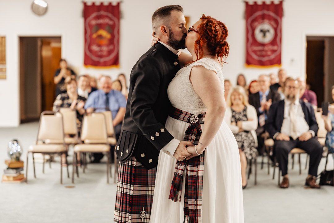 A romantic first kiss moment during a Scottish-themed wedding ceremony with guests in attendance.