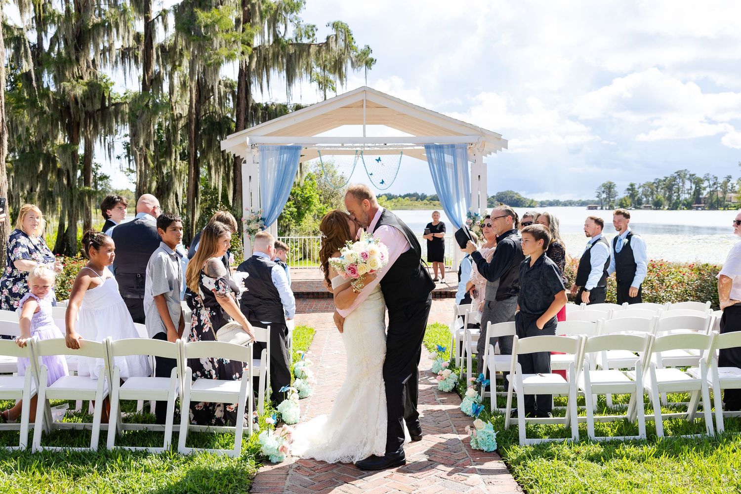 Newlyweds share first kiss as married couple while guests celebrate along brick ceremony aisle.