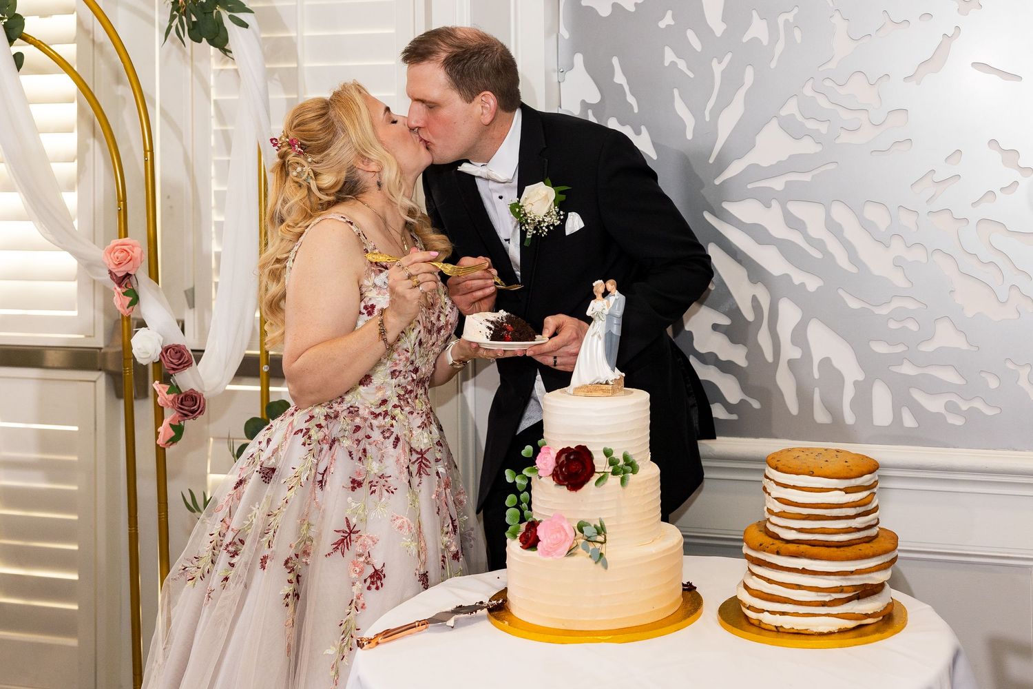 Bride and groom kiss while holding cake beside a decorated white wedding cake at the reception.