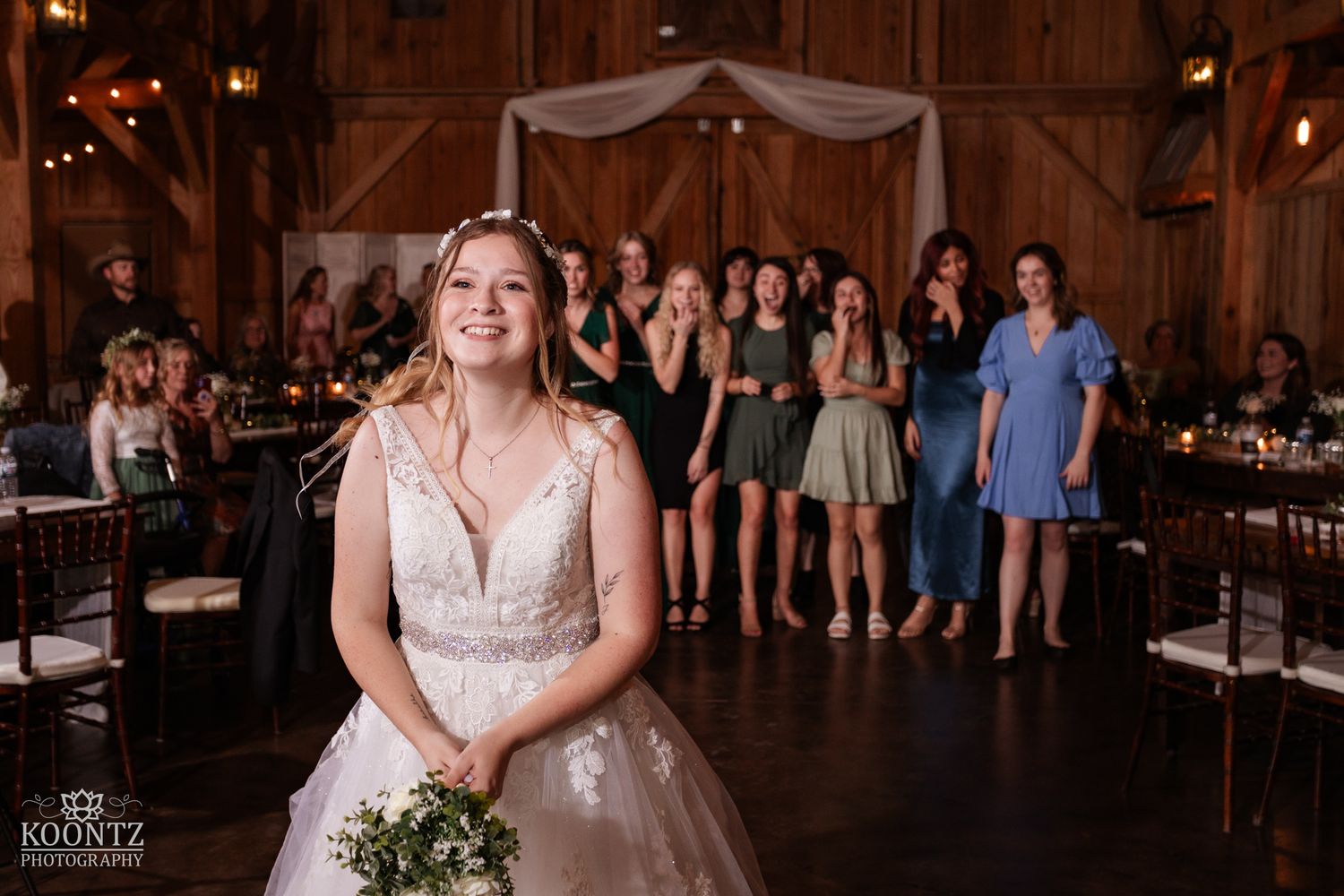Smiling bride prepares to toss bouquet to excited guests inside warmly lit rustic barn reception.