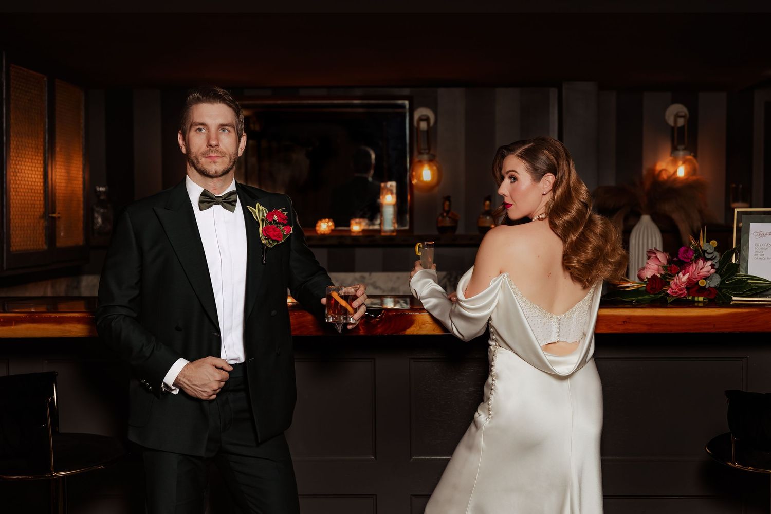 Stylish couple in wedding attire posing at a dimly lit bar.
