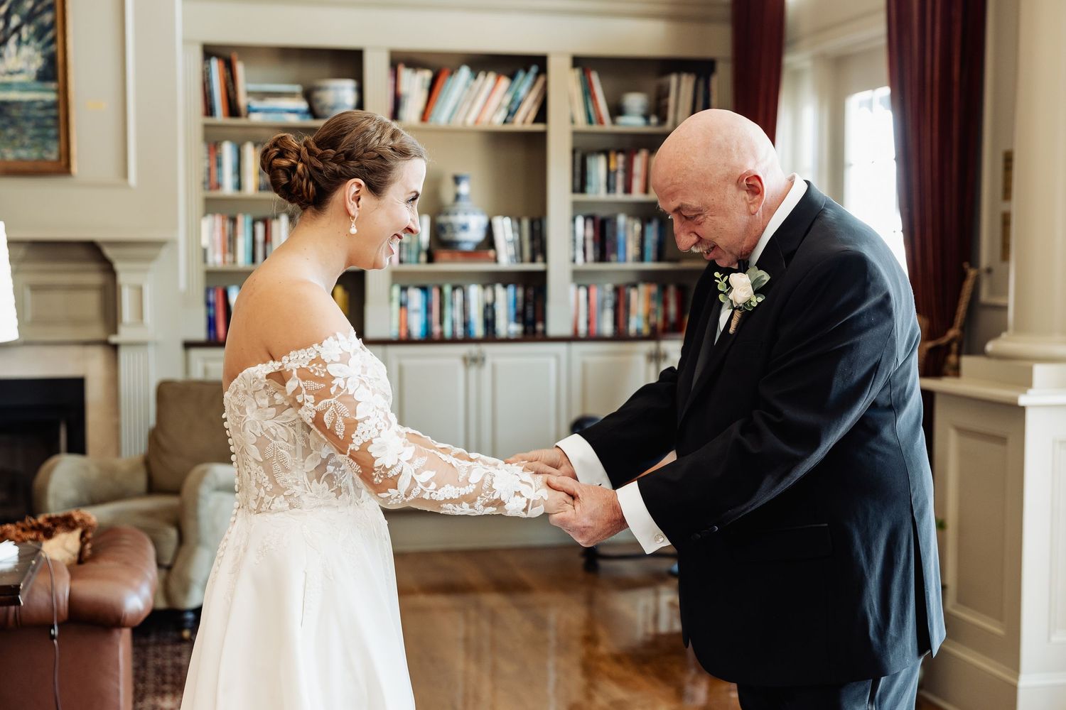 Bride and father share tender hand-holding moment during wedding first look in elegant library room.