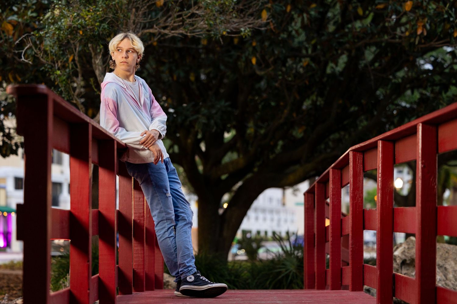 Teen boy in pink hoodie leans on red bridge railing in outdoor park setting.