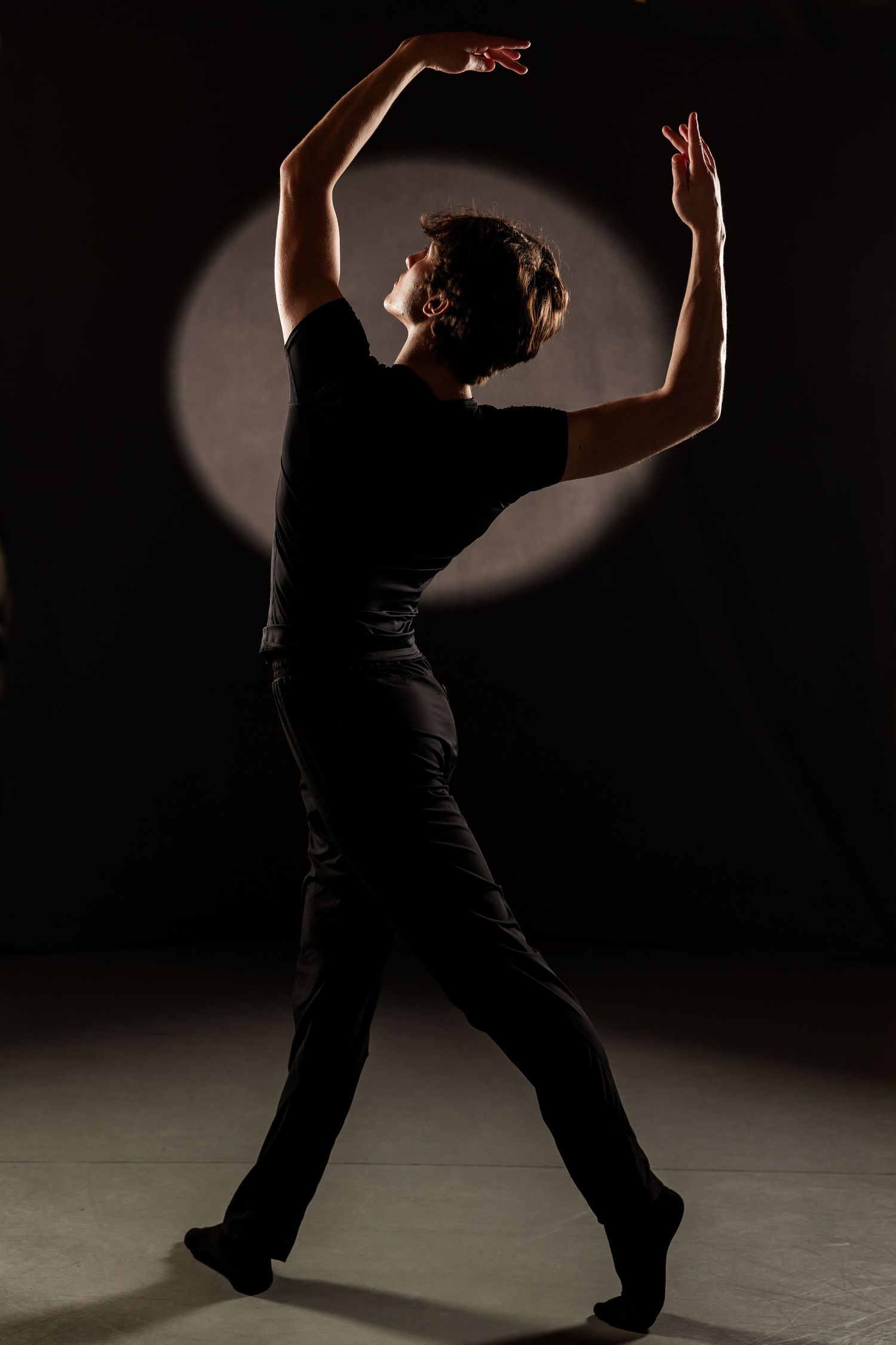 A dancer in black clothing performs an expressive pose with arms raised under a spotlight on a dark stage.