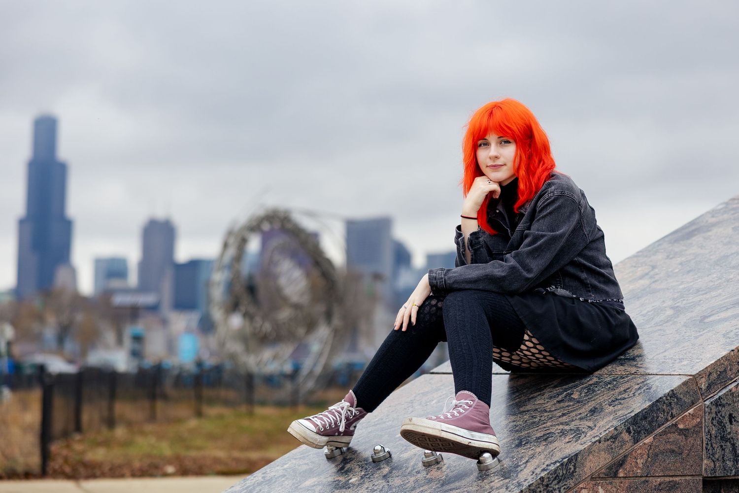 Young woman with bright orange hair sitting on a skateboard ramp with Chicago skyline in the background.