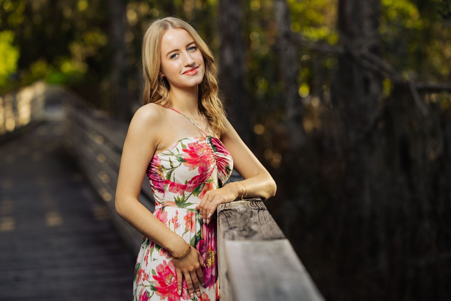 Young blonde woman in floral sundress smiling on wooden bridge surrounded by lush green trees.