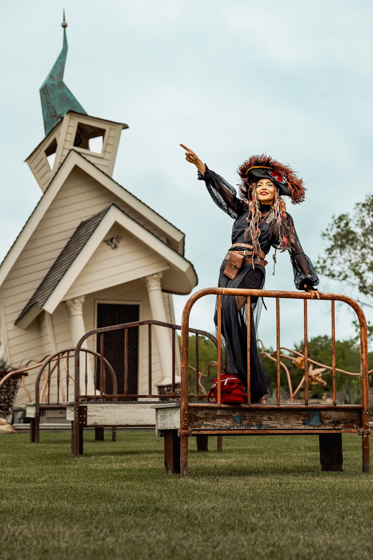 Woman in black pirate costume points from atop a rusty iron bed frame near white chapel.