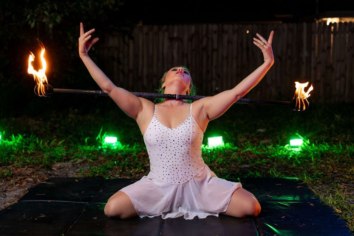 Female fire performer seated with arms raised holding a flaming staff during an outdoor night show.