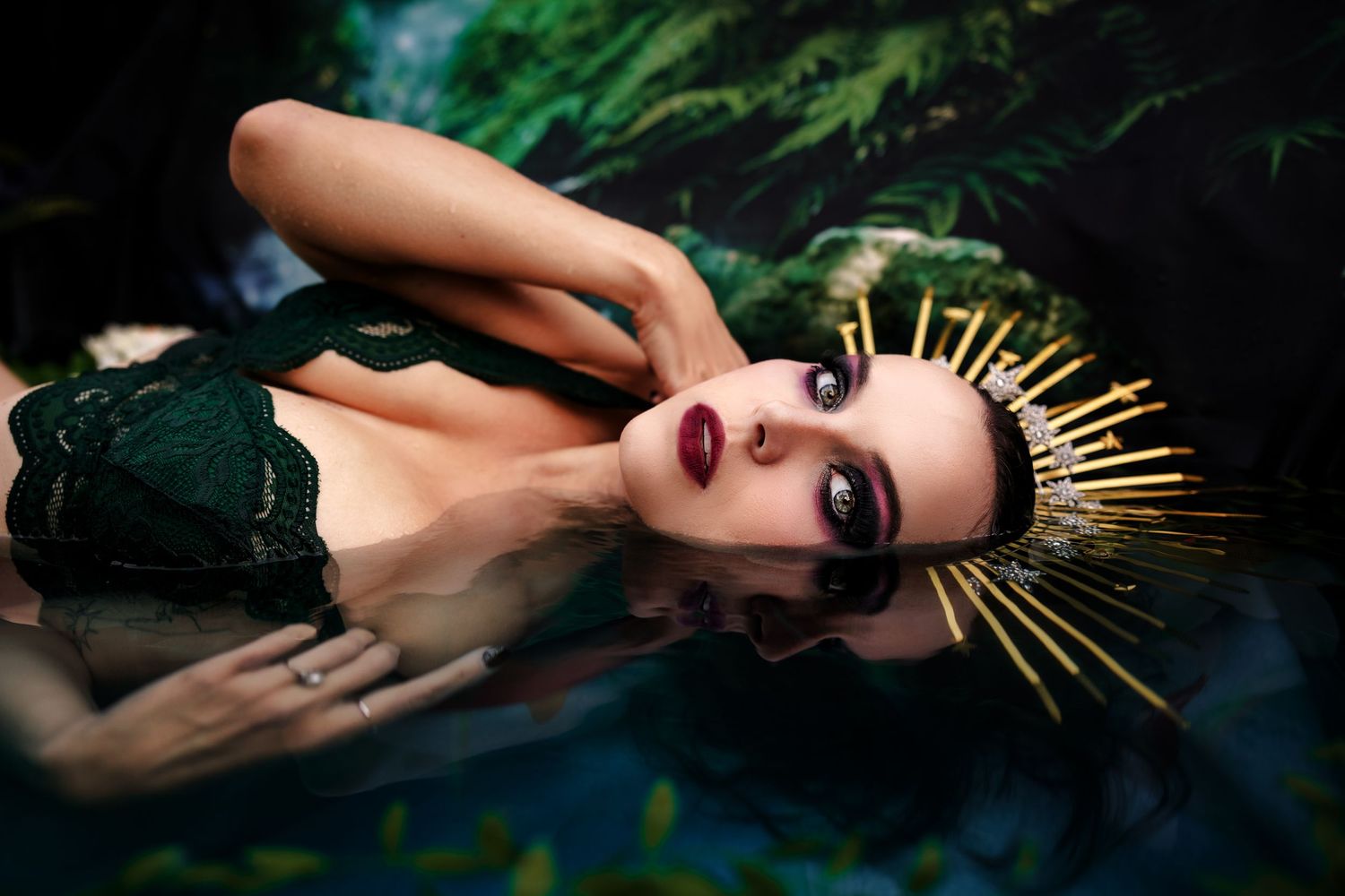 Woman in green lace bralette wearing gold sun crown lying in dark mystical water surrounded by foliage.
