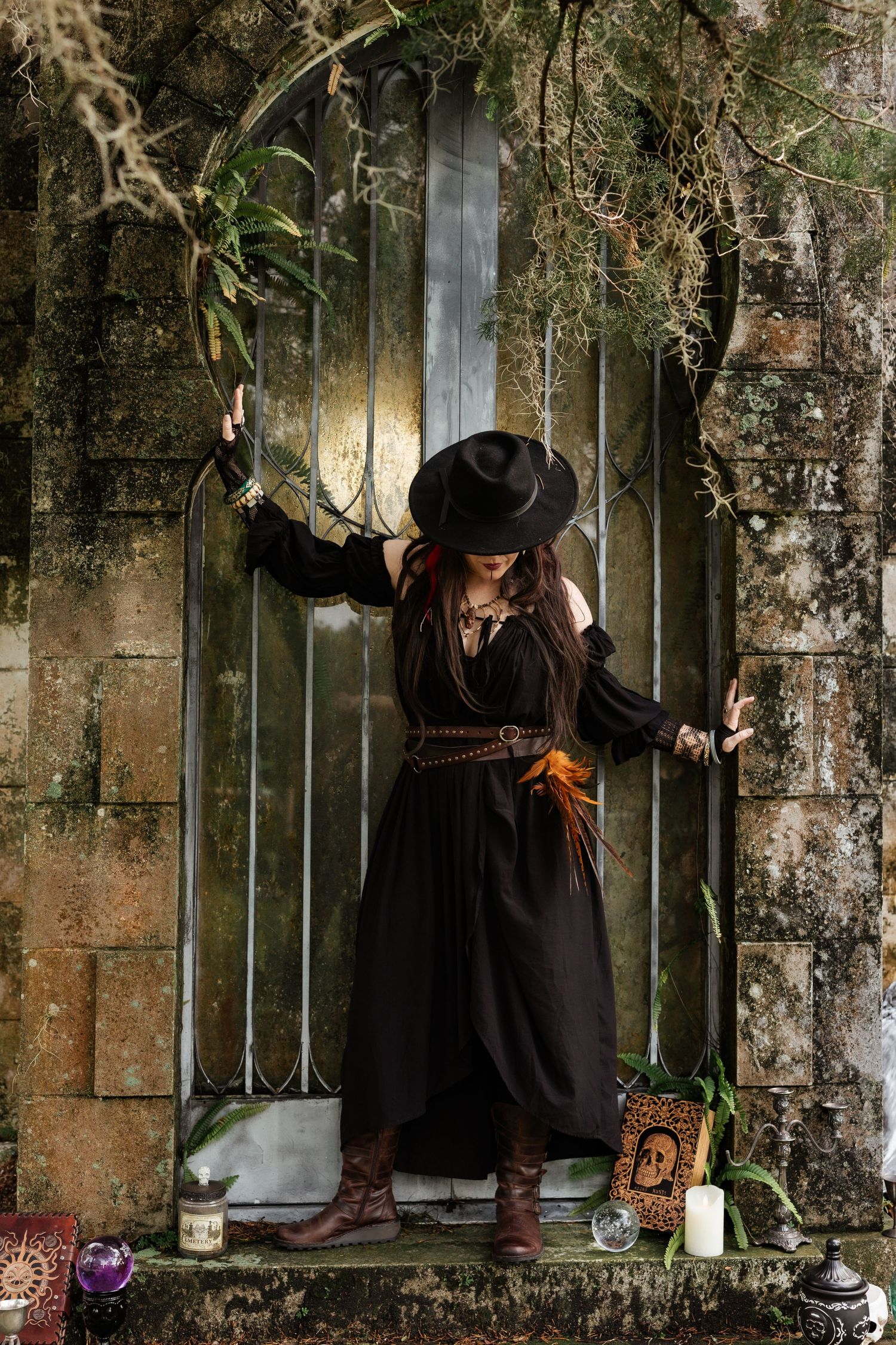 Witch in black dress and wide-brim hat stands at iron gate with candles, crystals, and skull book props.