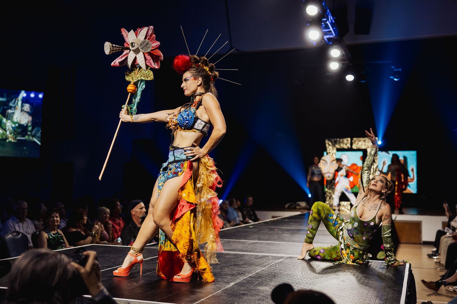 Model in vibrant fantasy costume with headdress walking a fashion show runway.