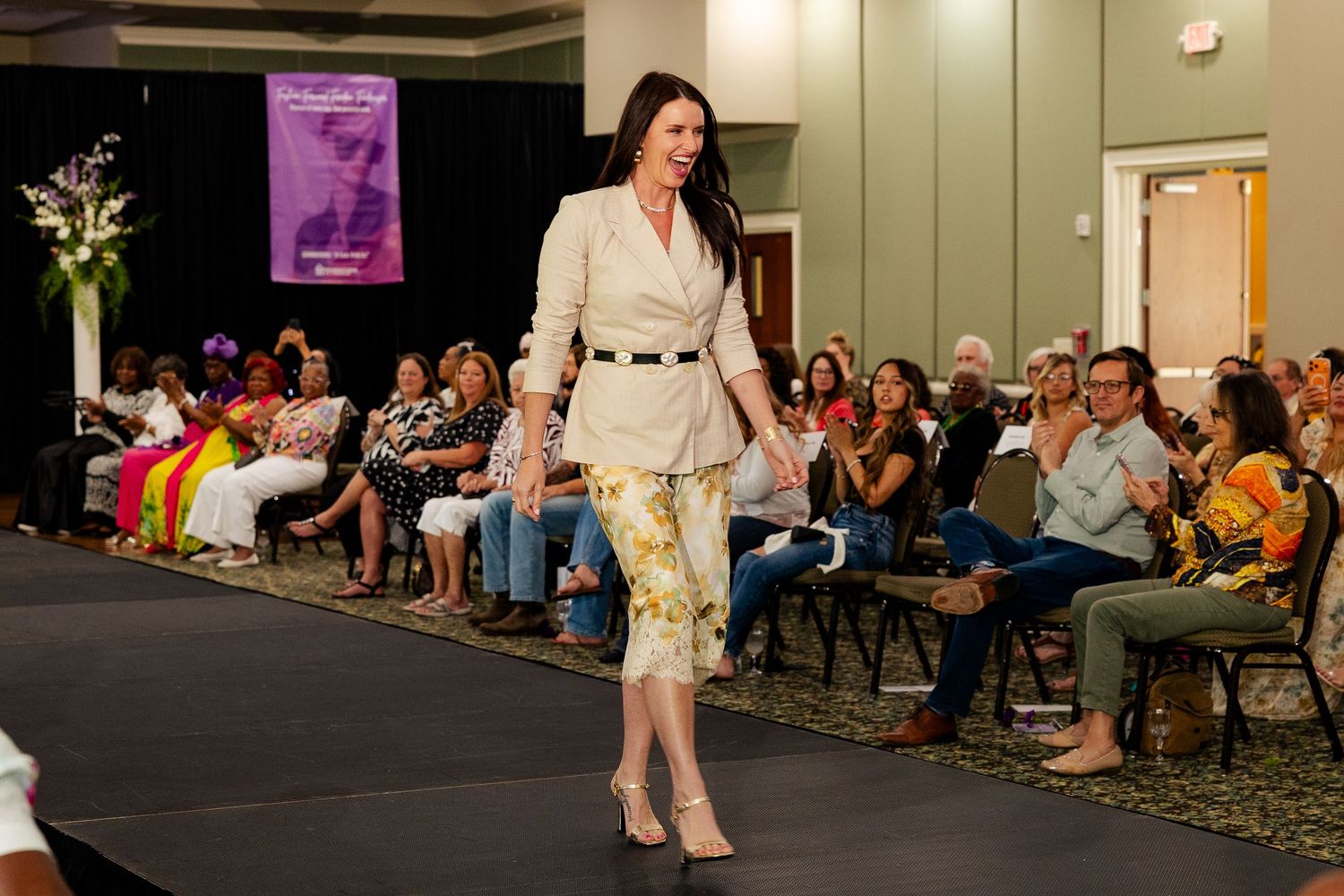 Woman models beige blazer and floral pants on runway at fashion show.