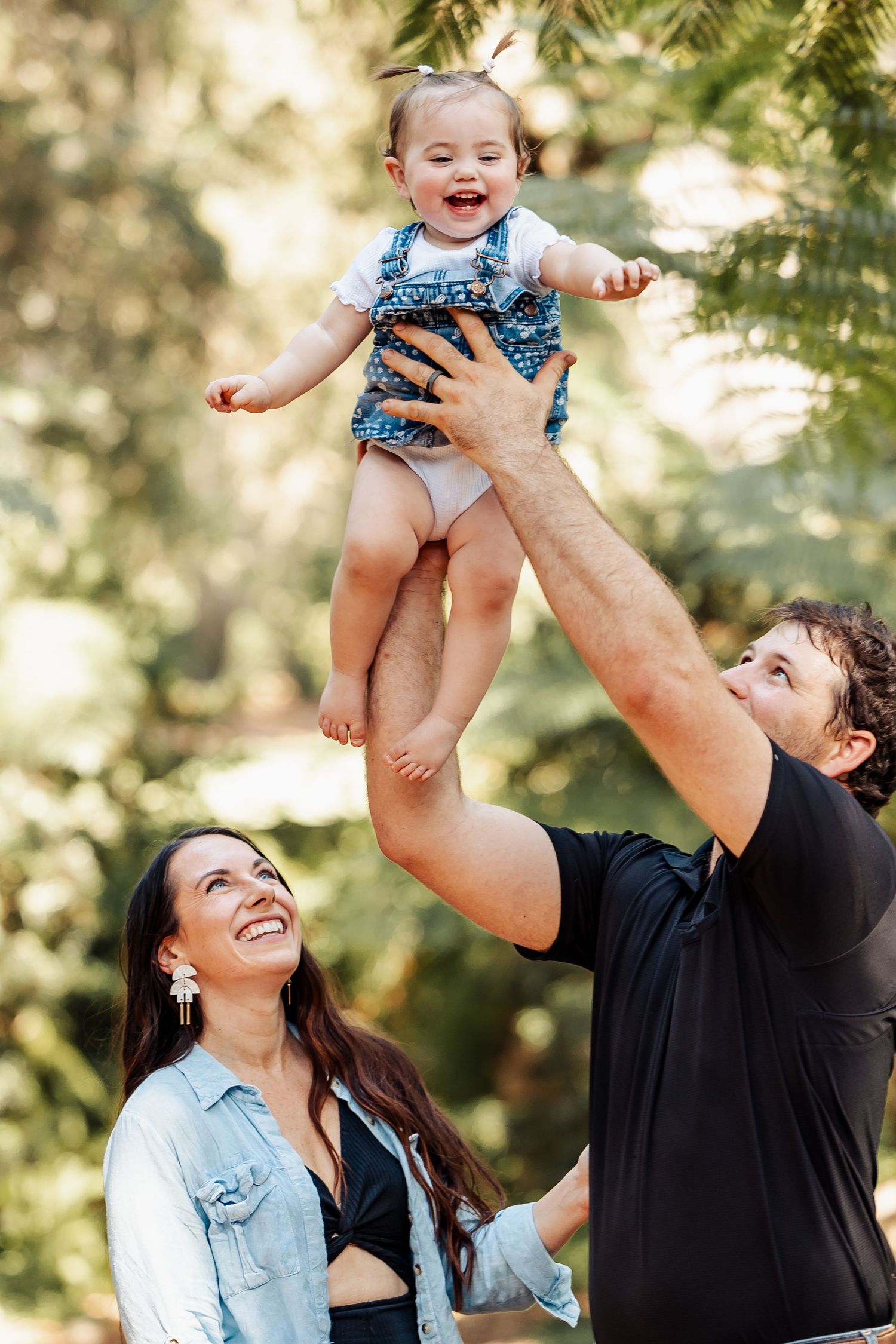 Joyful parents tossing laughing baby girl in the air outdoors among green trees.