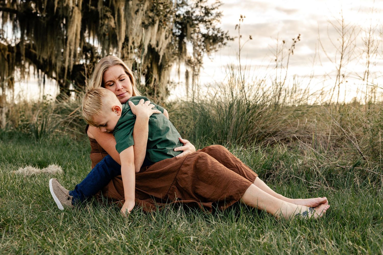 A mother sits in lush green grass hugging her young child outdoors at golden hour near moss-draped trees.
