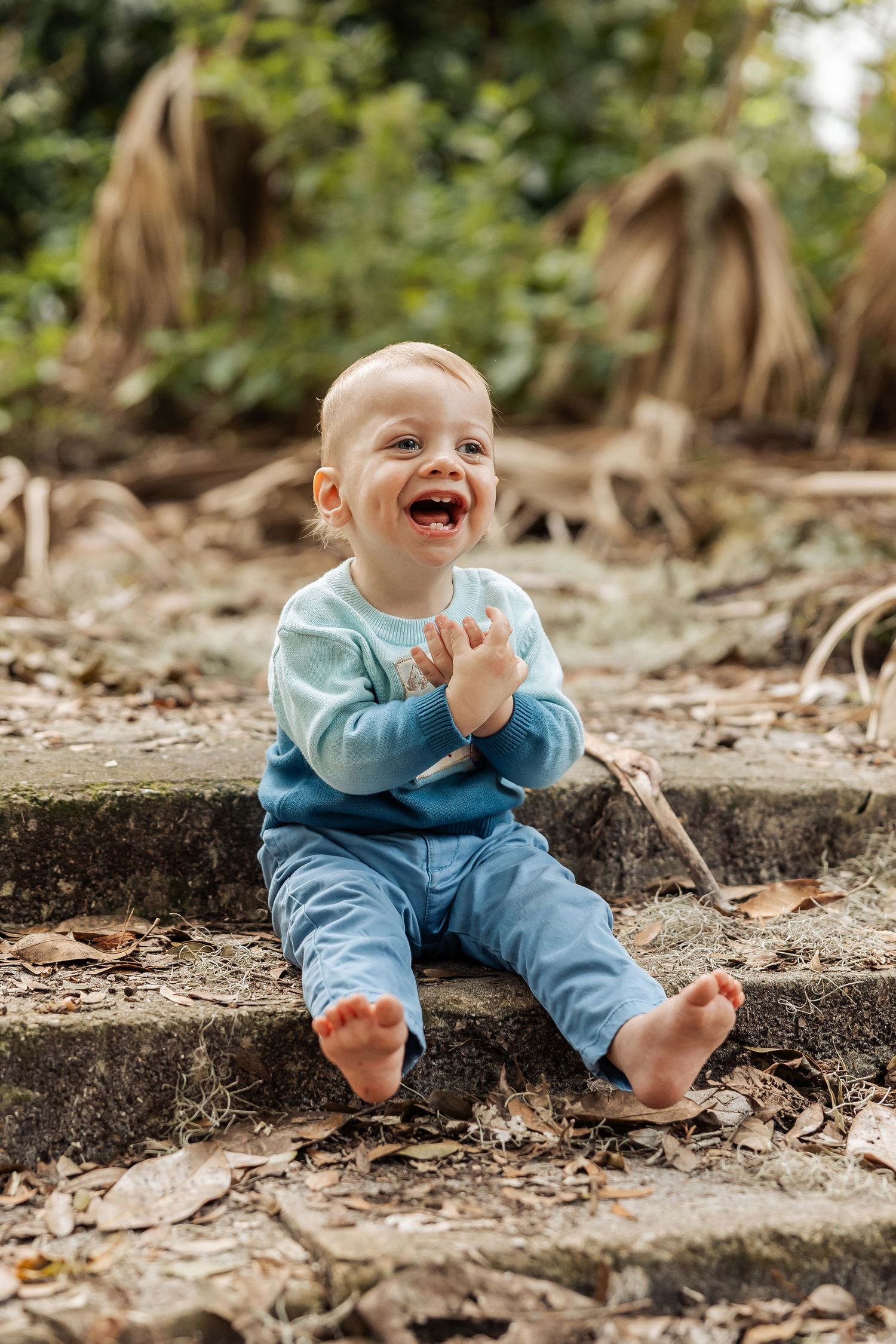 A happy baby in a blue outfit sits barefoot on stone steps laughing and clapping outdoors.