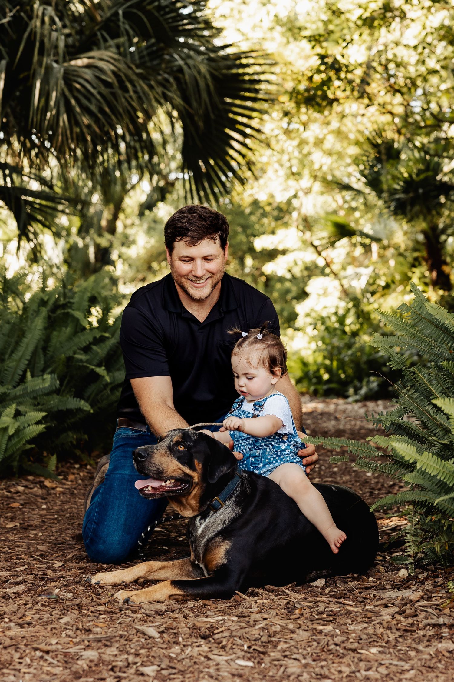 Smiling father kneels with baby girl sitting on their Rottweiler dog in a sunny garden.