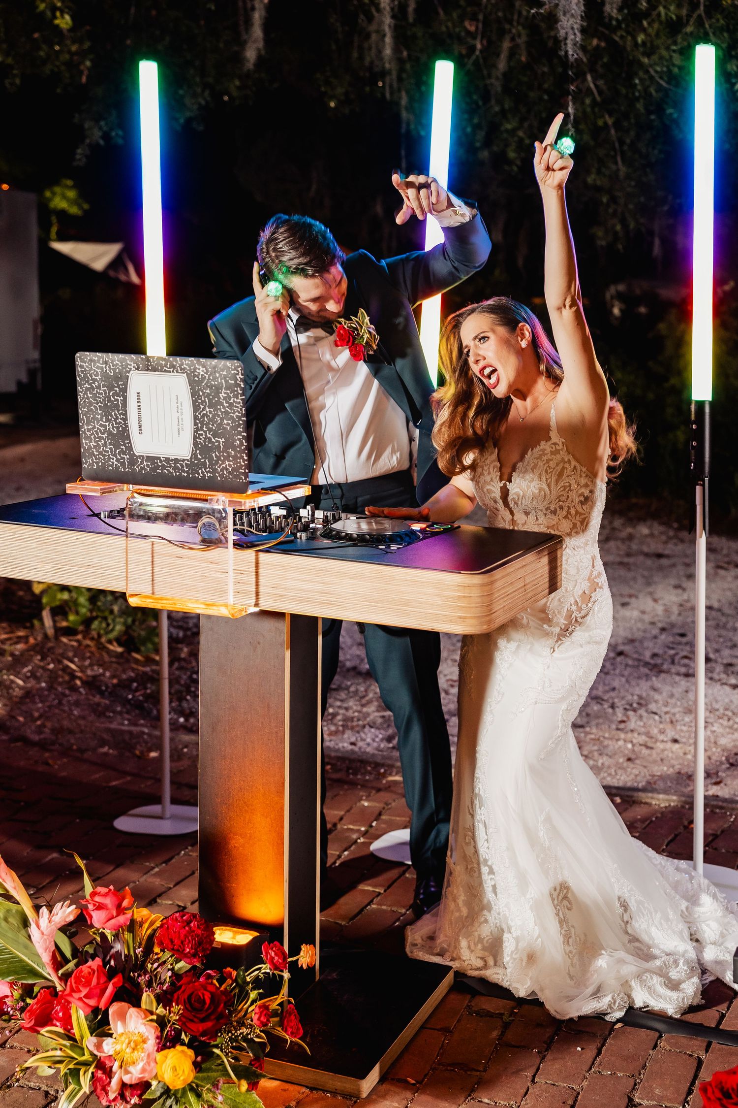 Bride and groom celebrating at DJ booth with colorful neon lights at night.