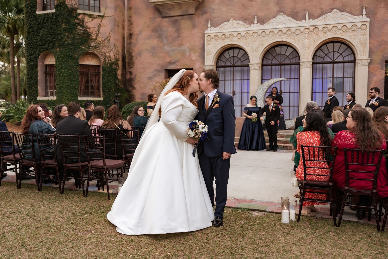 Newlyweds share romantic kiss at end of aisle after outdoor wedding ceremony at historic venue.