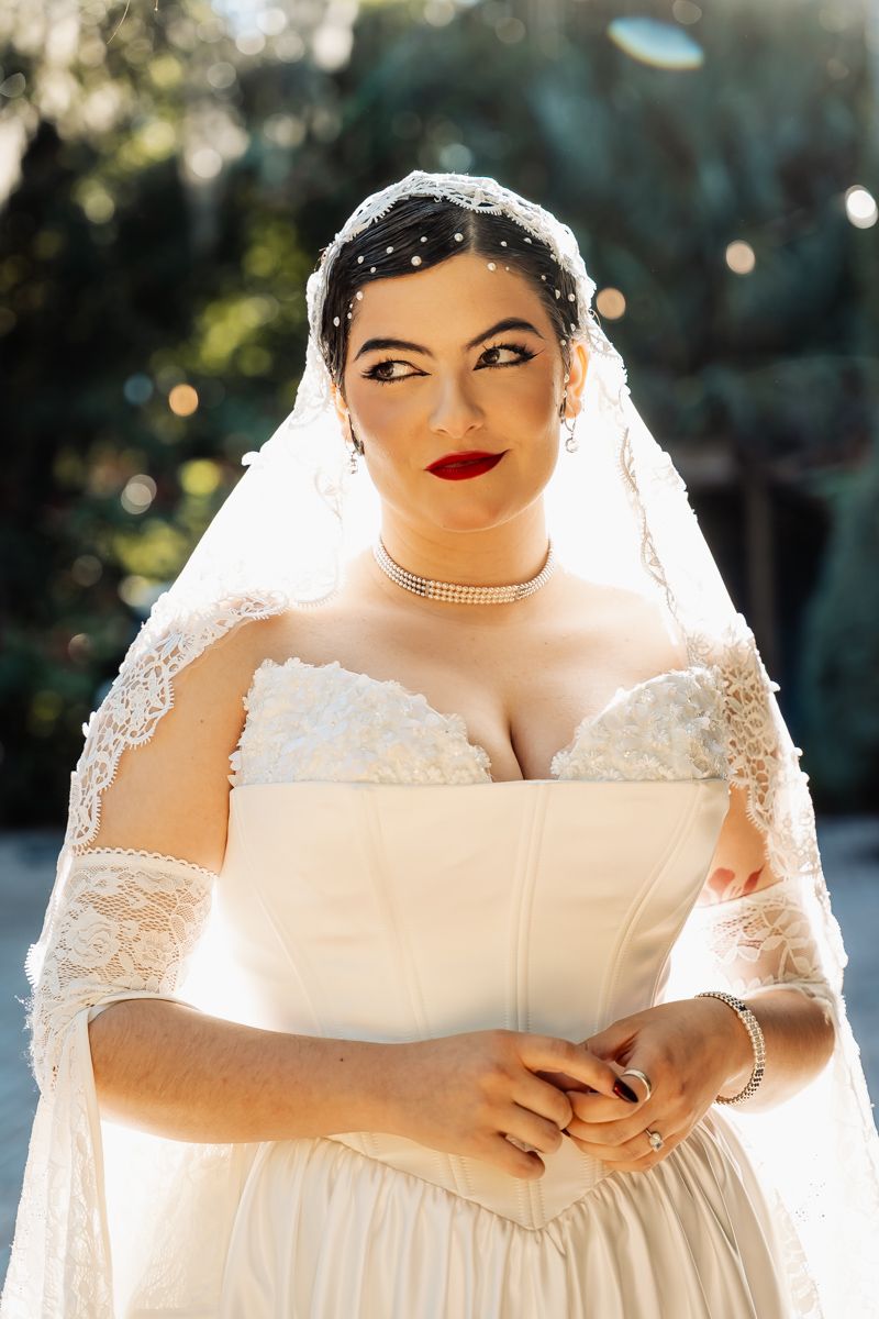 Elegant bride in off-shoulder lace wedding gown with pearl veil and red lips posing outdoors.