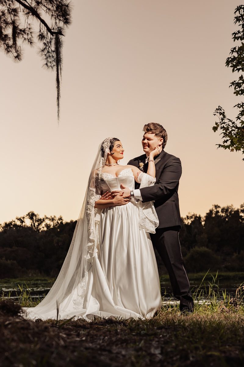 Bride and groom share romantic sunset portrait outdoors near lake, bride touching grooms face.