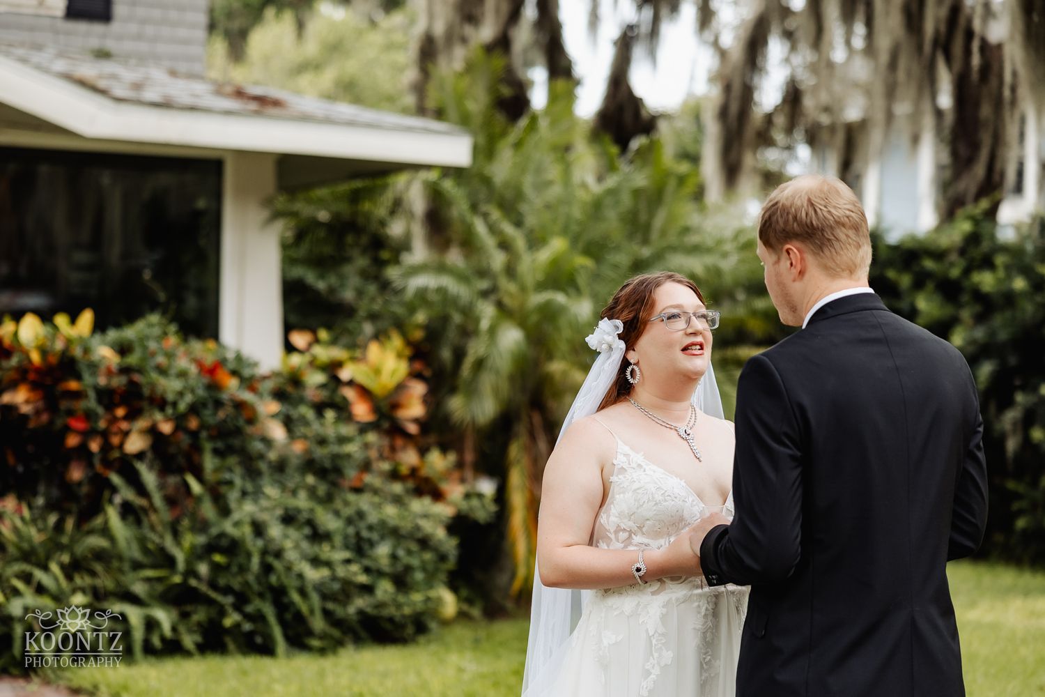 A bride and groom share a sweet moment in front of a southern home with lush landscaping and Spanish moss.