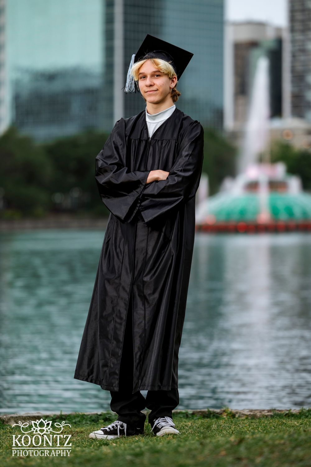 Graduate in black cap and gown poses by urban fountain with cityscape backdrop.
