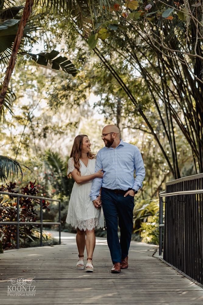 A romantic couple walks along a wooden boardwalk surrounded by lush bamboo and tropical foliage.
