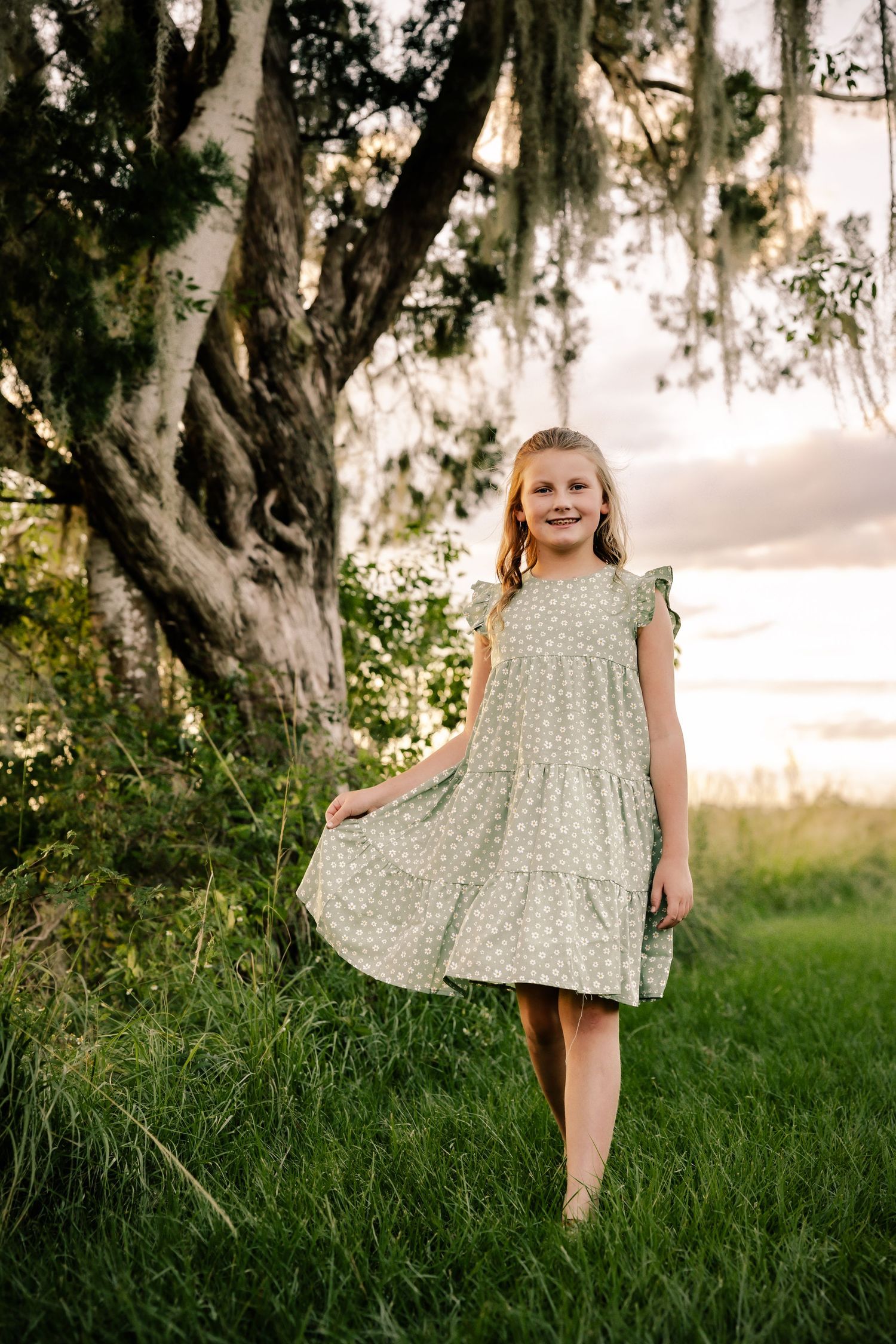 A young girl in a floral dress twirls outdoors under a tree at sunset.