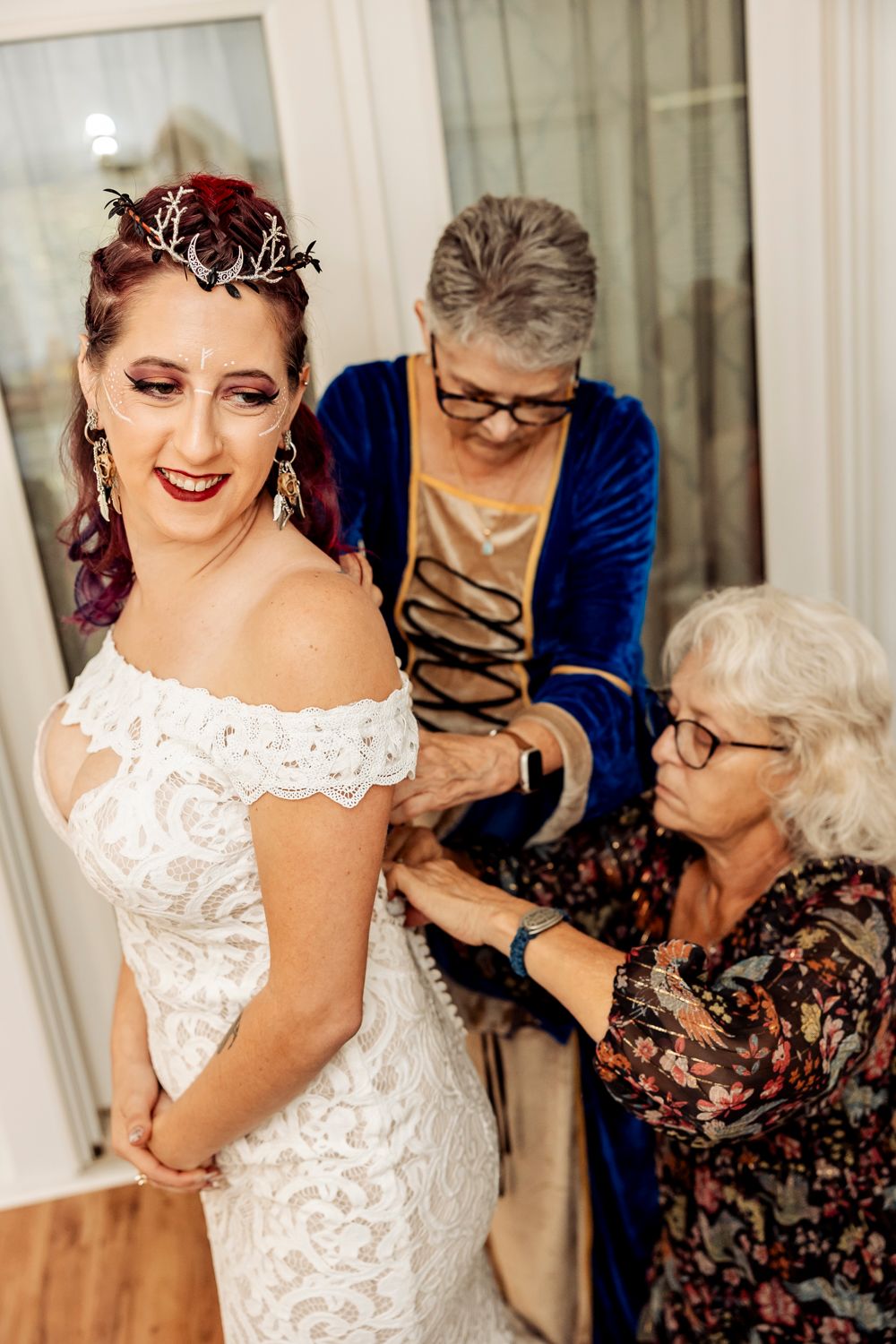 Bride in lace off-shoulder gown gets help fastening dress from two older women before her wedding ceremony.
