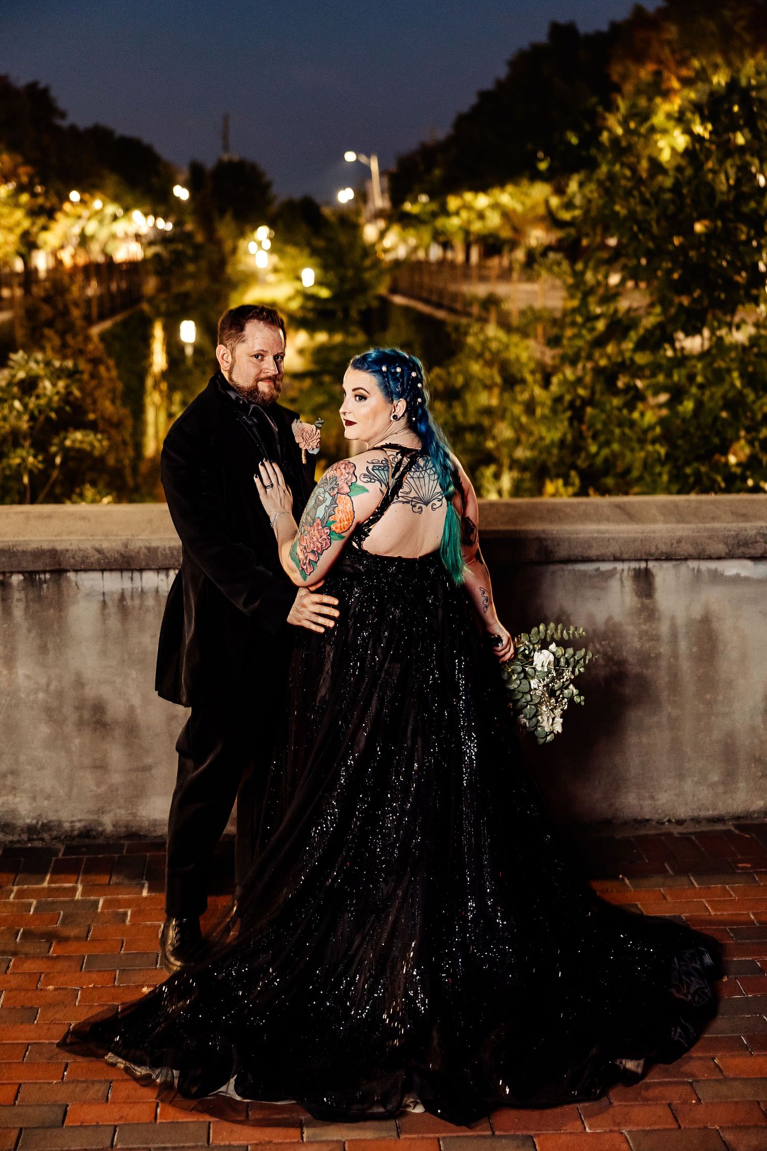 Bride in backless black sequin gown with teal hair and groom pose outdoors at night.