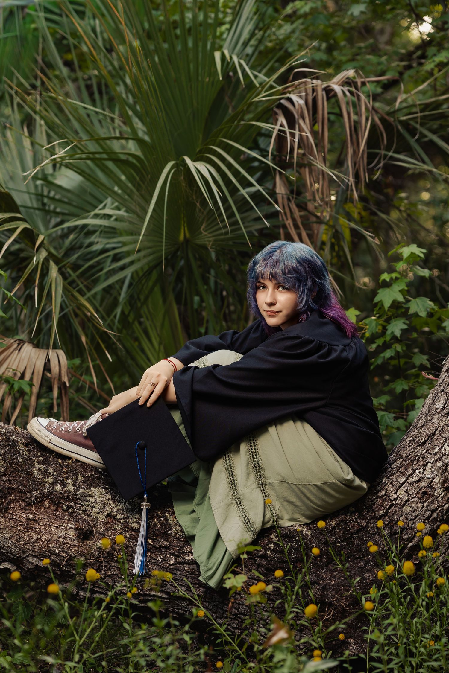 Blue-purple haired graduate in black gown sits against tree trunk holding graduation cap outdoors.