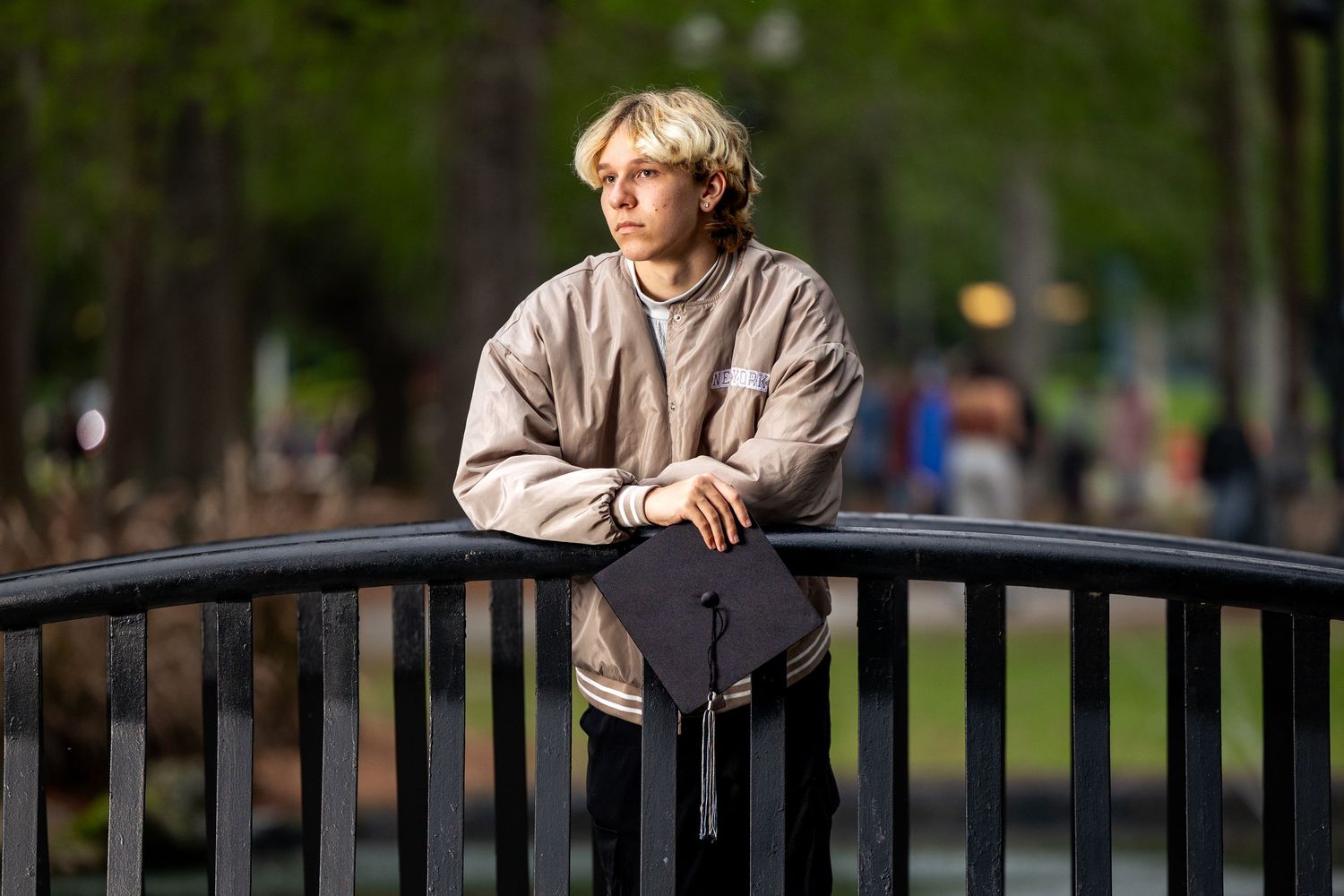 Blonde teenage boy leaning on black metal railing in sunny park, wearing beige jacket with arms crossed.