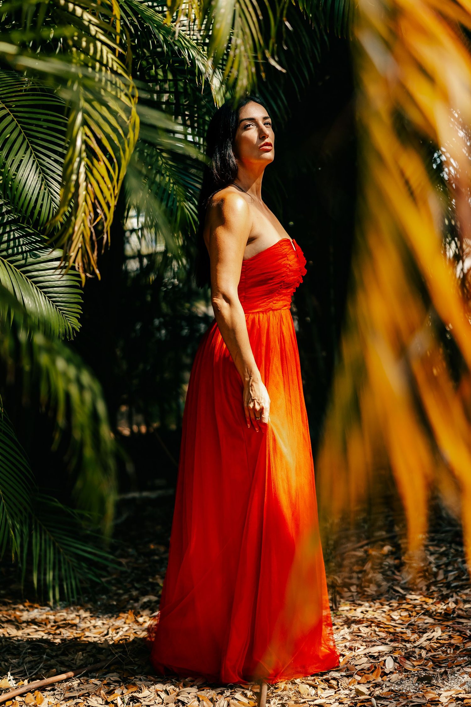 Woman in red strapless gown standing among tropical palm trees in golden sunlight.