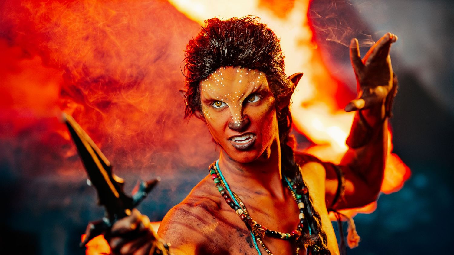 Close-up of fierce fantasy demon cosplayer with pointed ears, tribal makeup, and fire background.