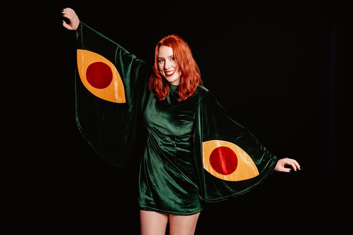 Smiling redhead woman in green velvet avocado Halloween costume poses against black background.