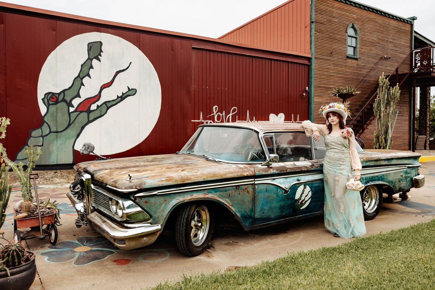 Woman in vintage mint gown poses beside a rustic patina classic car near a colorful mural.