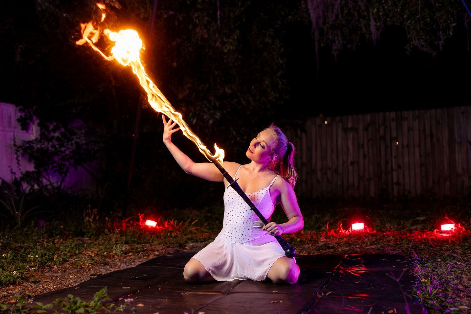 Female fire artist seated on ground extending a blazing fire staff upward during nighttime performance.