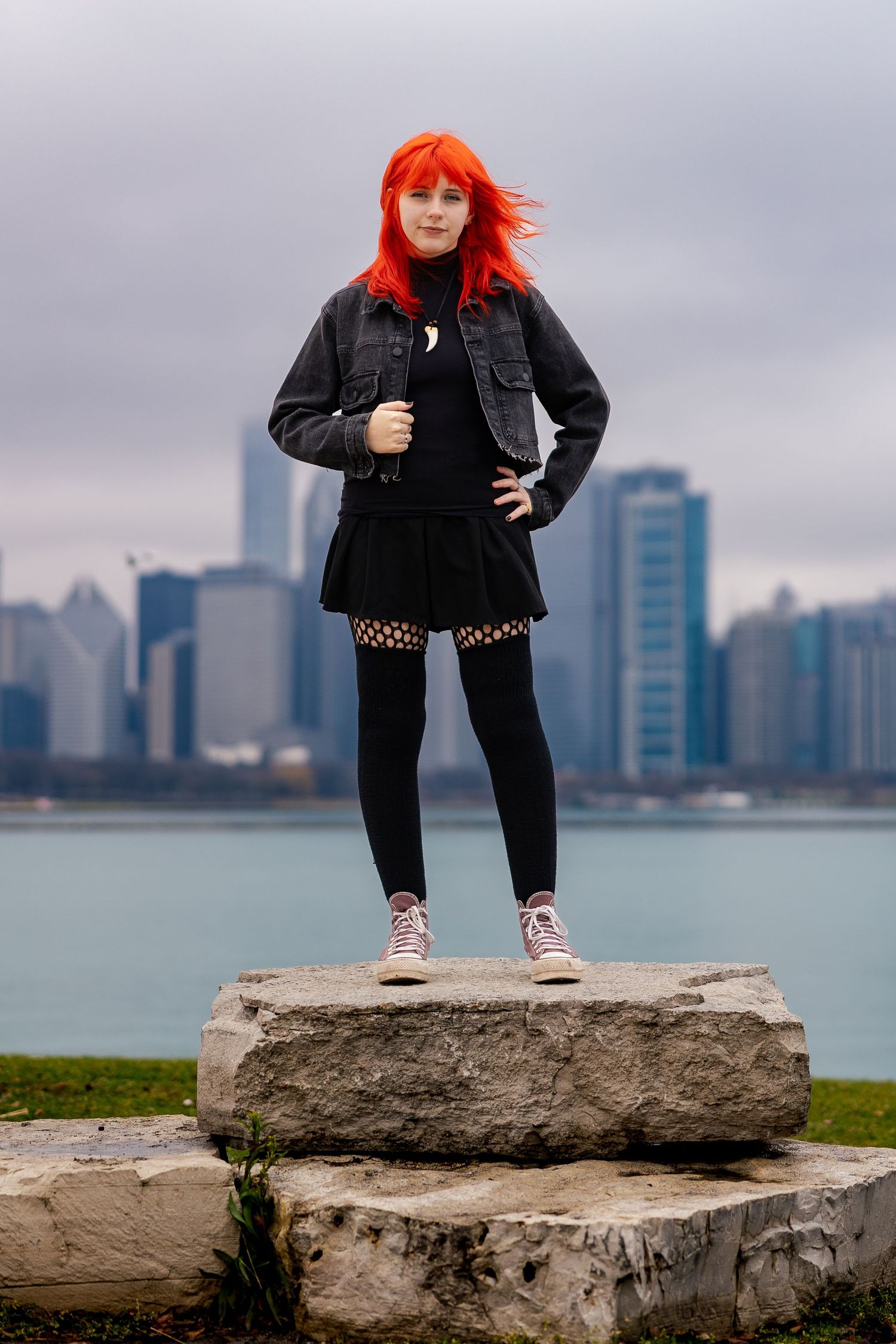 Young woman with bright red hair in black outfit stands on rocks with city skyline and lake in background.