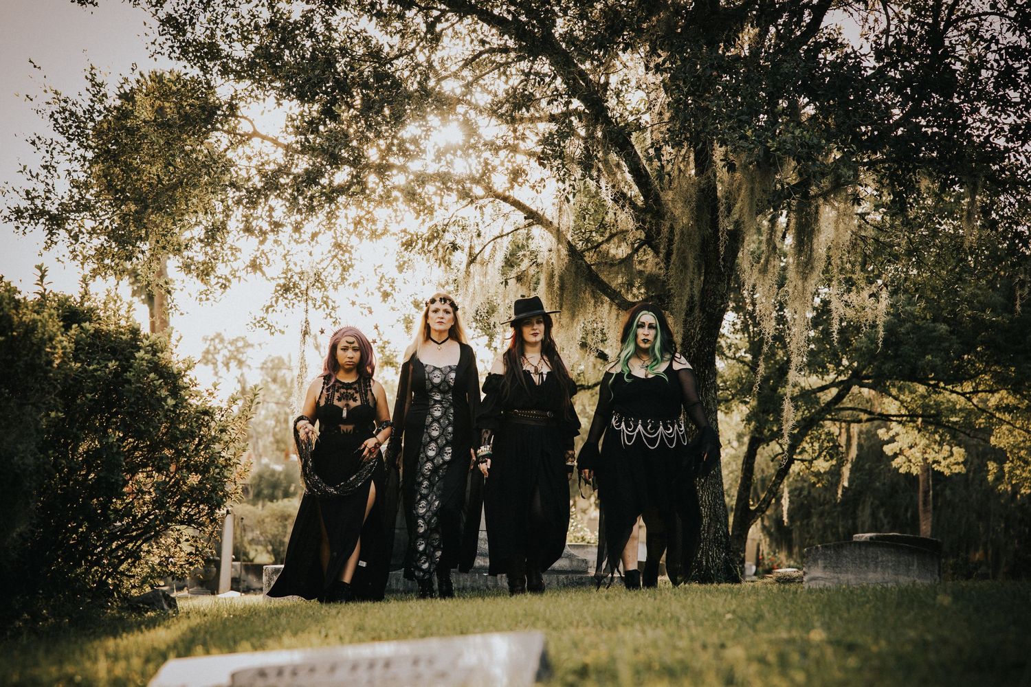 Four women dressed in black witch costumes stand together in a sunlit cemetery with moss-draped trees.