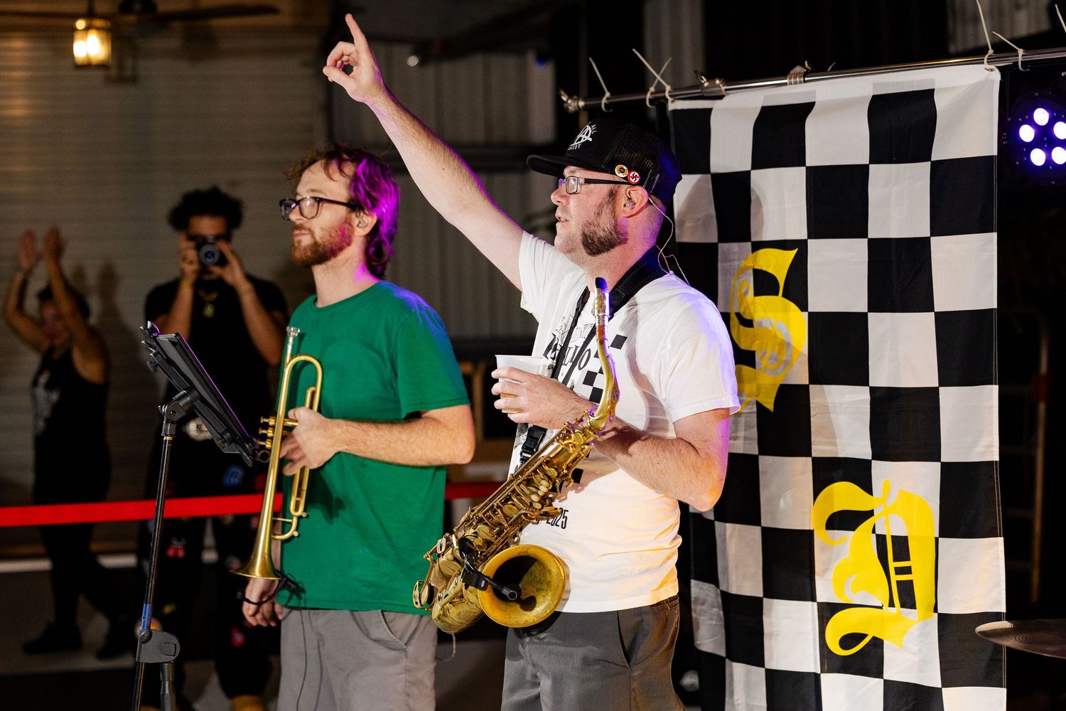 Two musicians perform on stage, one playing trumpet and one playing saxophone, with a checkered flag backdrop.