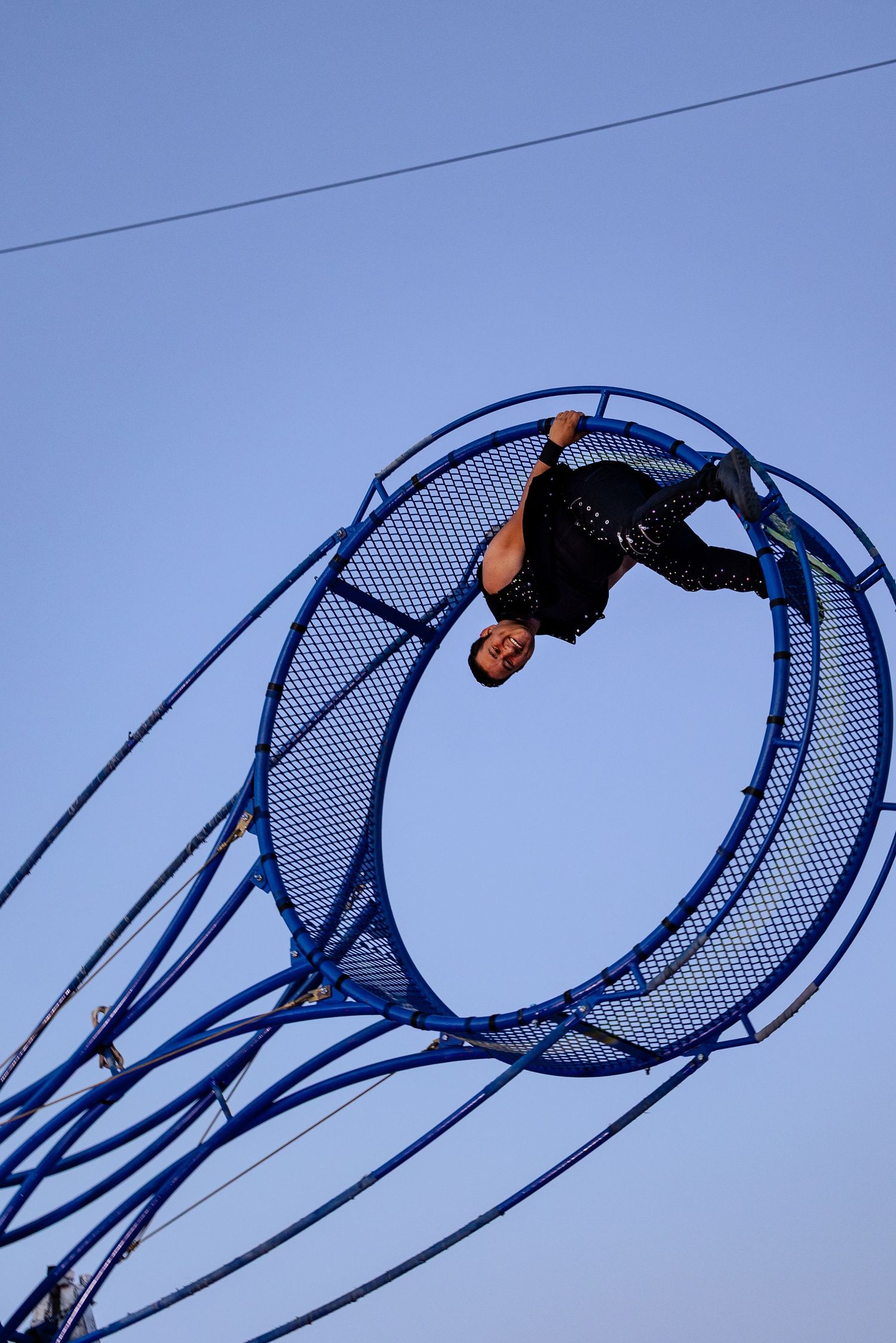 Acrobat performing daring aerial stunt inside a large blue circular wheel apparatus against a clear blue sky.