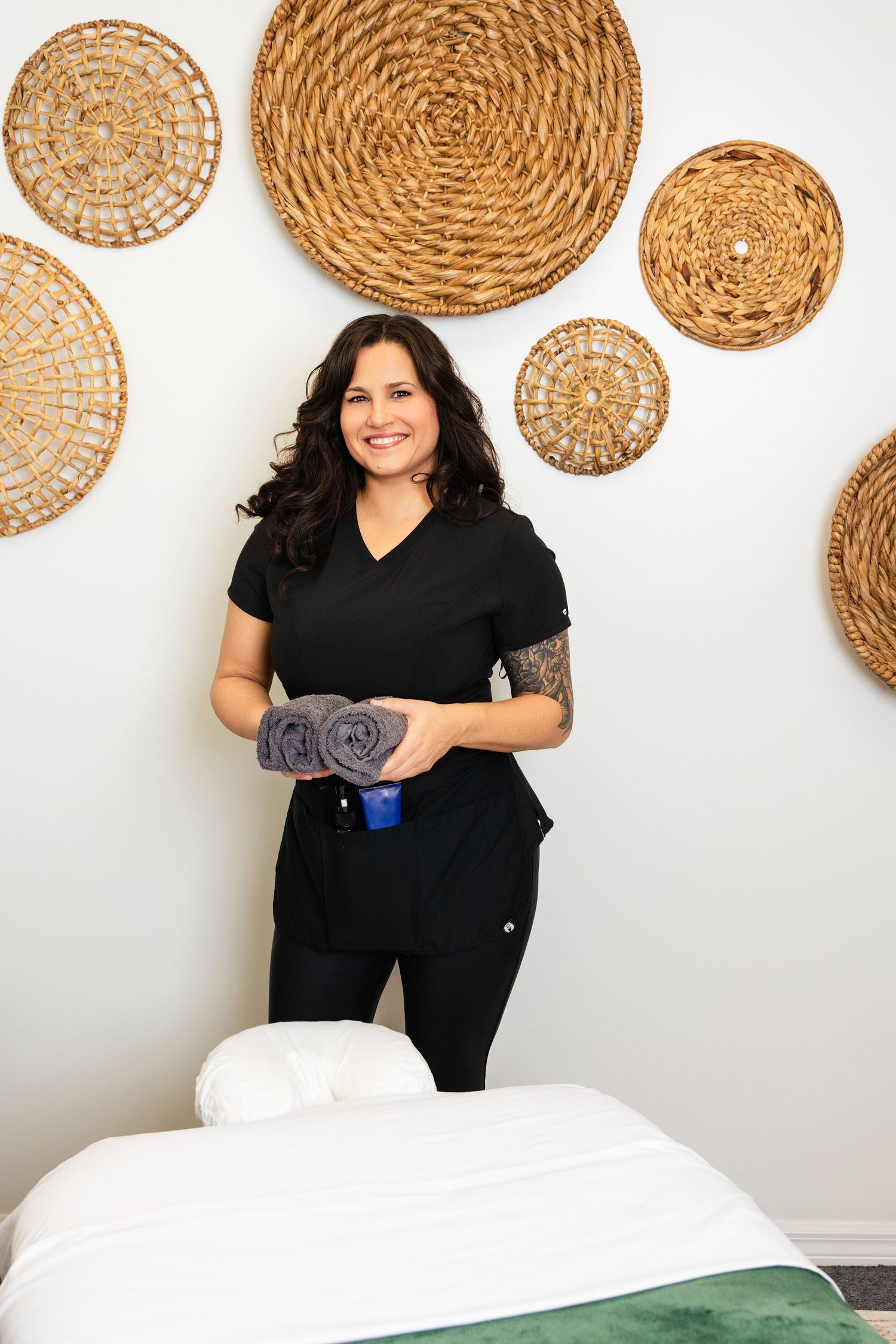 Female massage therapist in black scrubs holding rolled towels beside a massage table in a spa.