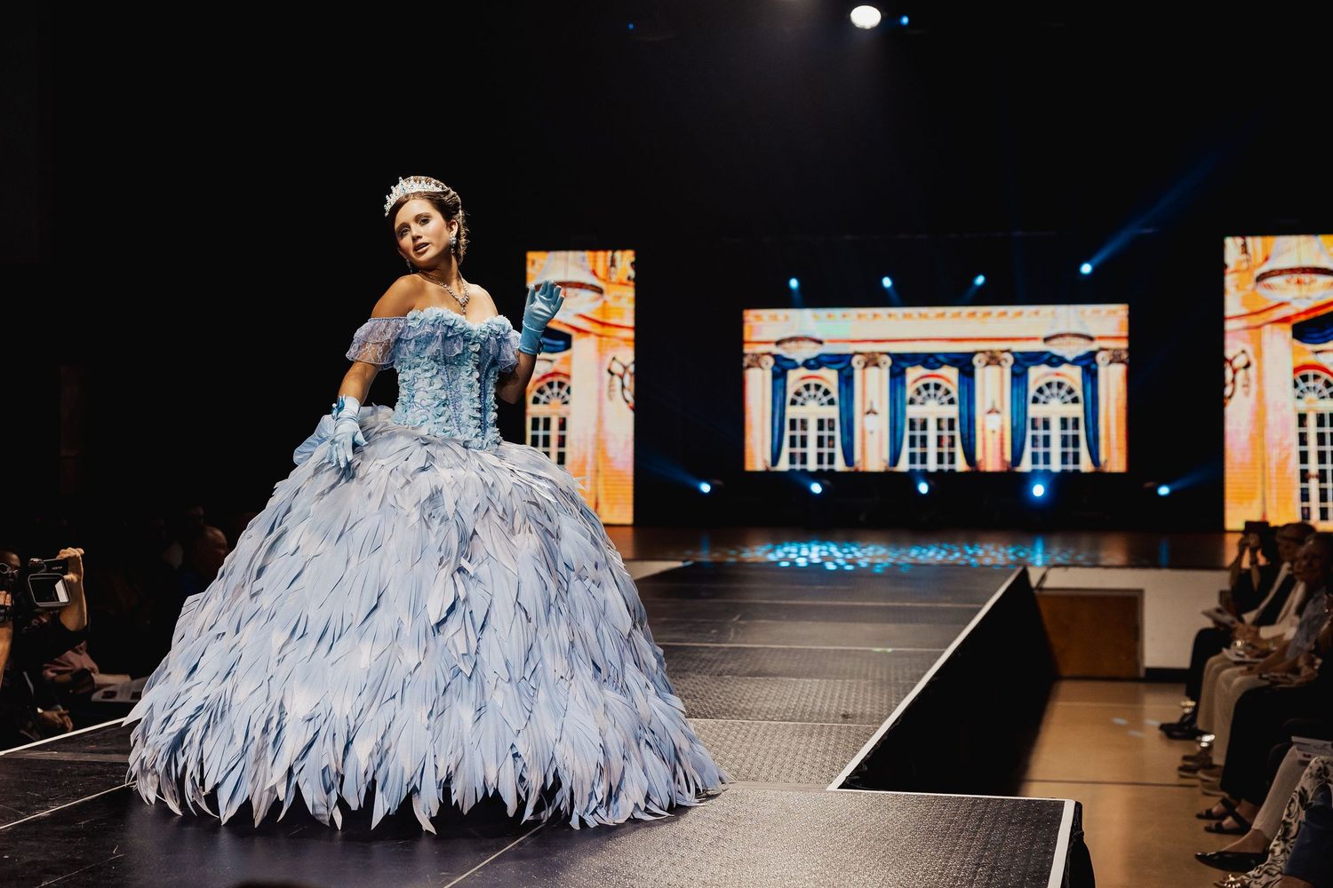 Model in an elaborate blue feathered ball gown and tiara on a fashion runway.