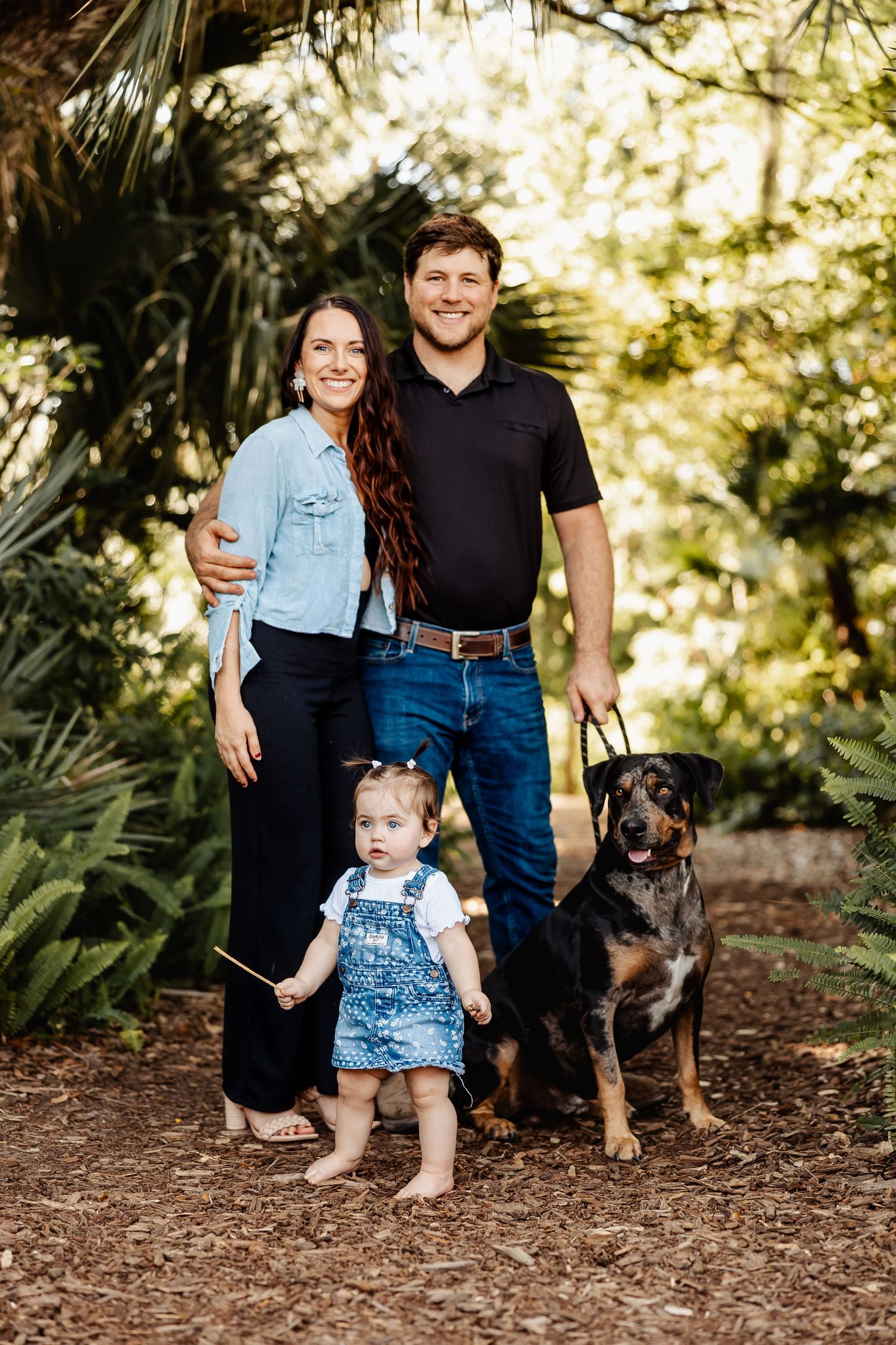 Smiling family with a baby girl and Rottweiler dog pose together in a lush green garden setting.