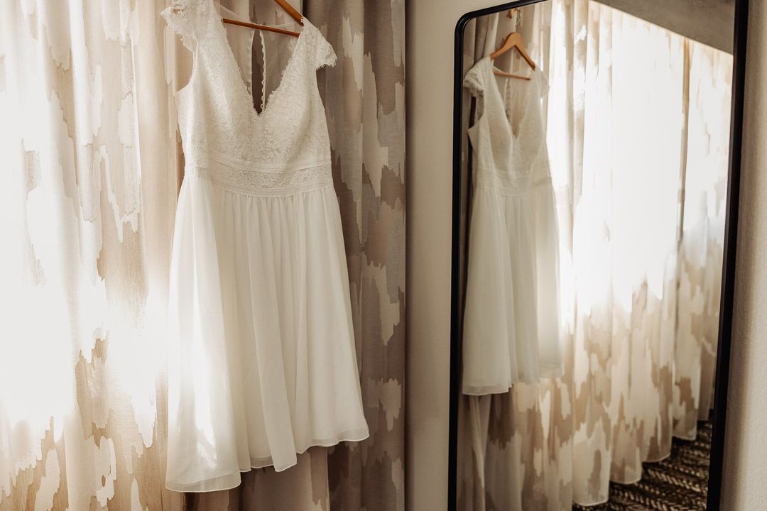 White lace wedding dress hanging on a hanger reflected in a full-length mirror in a bright room.