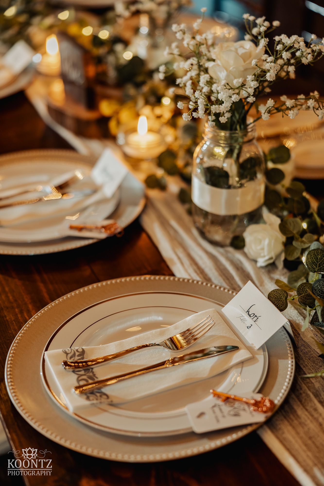 Elegant rustic wedding table setting with gold cutlery, white roses, and candlelit centerpieces.