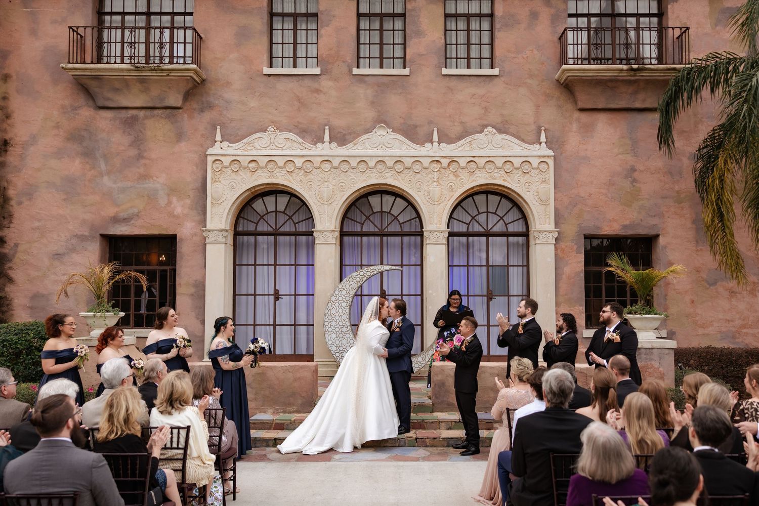 Bride and groom kiss at outdoor ceremony altar before Spanish-style mansion with arched windows.