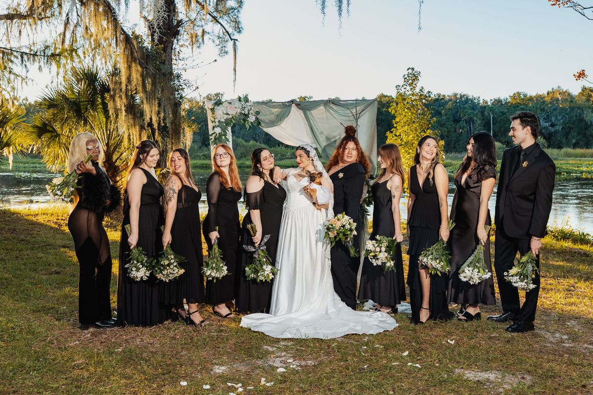 Bride with bridesmaids in black dresses holding greenery bouquets pose near lakeside ceremony arch.
