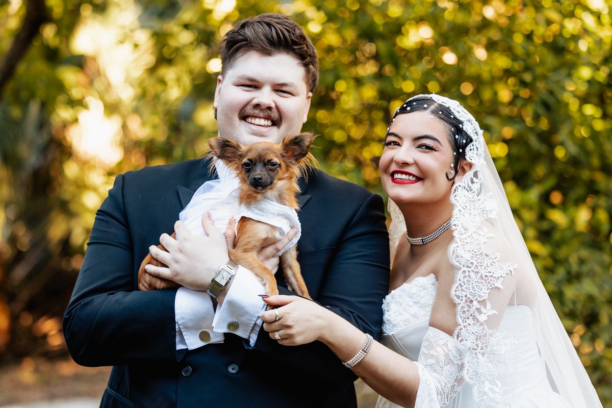 Smiling bride and groom holding small dog dressed in wedding attire outdoors in autumn setting.