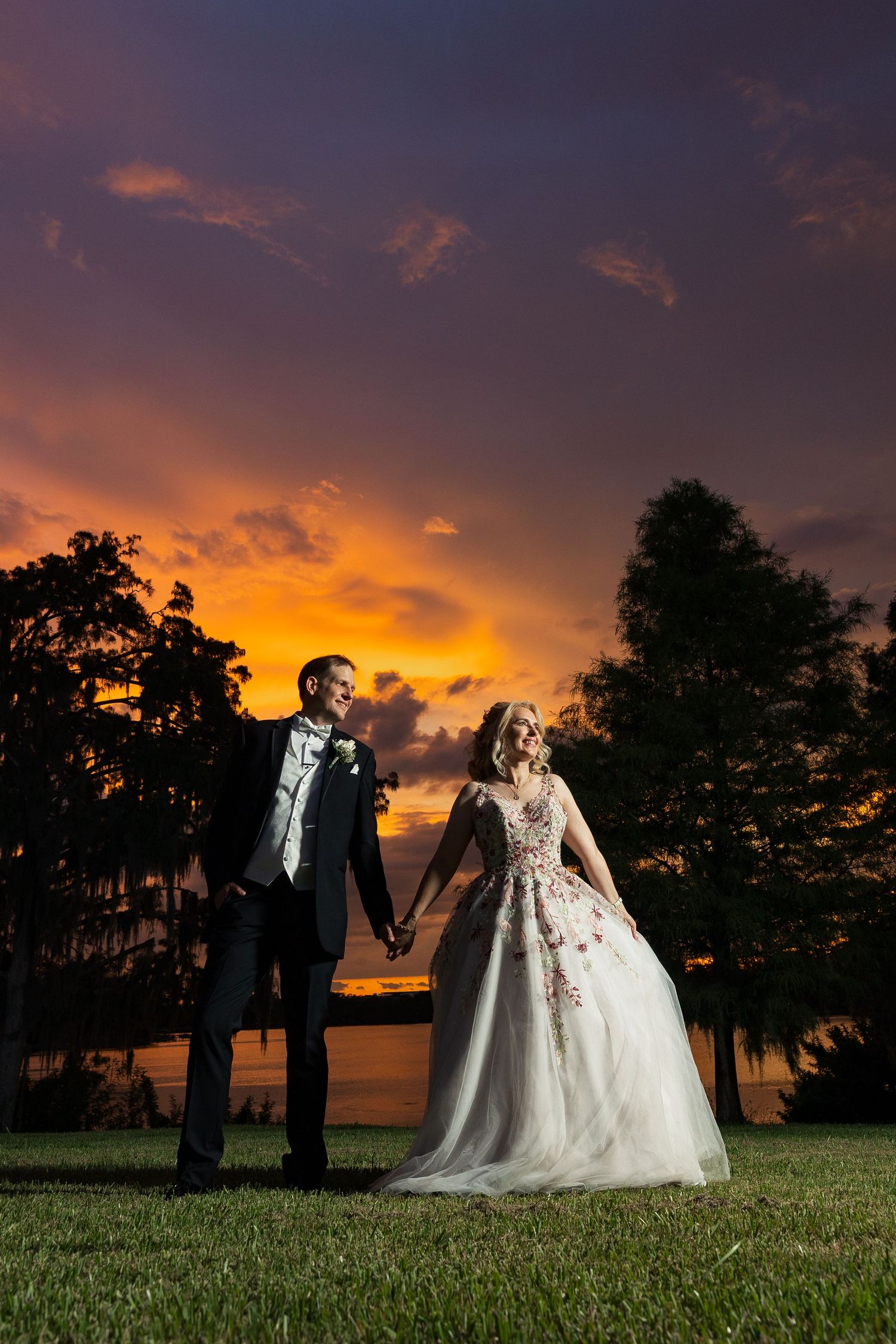 Newlyweds hold hands on a lawn with silhouetted trees and a vibrant orange and purple sunset.